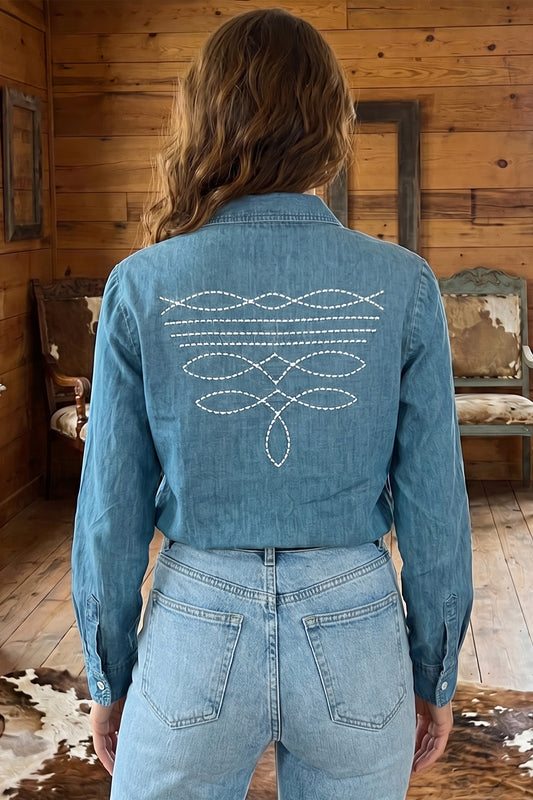 A woman viewed from behind wearing a blue denim shirt with white embroidered geometric patterns on the back, paired with light-wash jeans in a rustic wood-paneled room.
