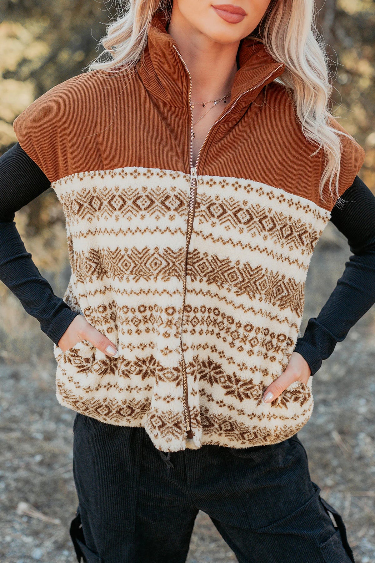 A woman wearing a brown and cream patterned fleece vest over a black long-sleeve top and black pants, photographed outdoors with a blurred natural background.