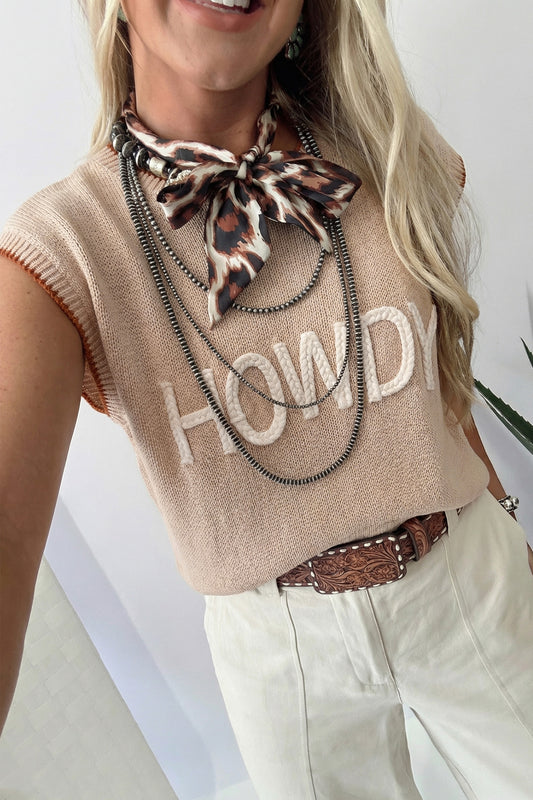 Person wearing a beige sleeveless sweater with 'HOWDY' text, leopard print scarf, and white pants.