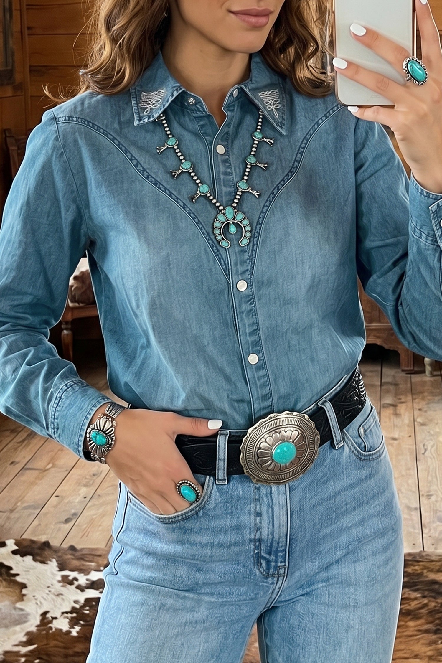 A woman wearing a light blue denim western-style button-up shirt with embroidered yoke detailing, paired with jeans, a black belt, and turquoise statement jewelry, shown in a mirror selfie.