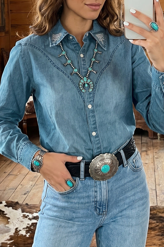 A woman wearing a light blue denim western-style button-up shirt with embroidered yoke detailing, paired with jeans, a black belt, and turquoise statement jewelry, shown in a mirror selfie.