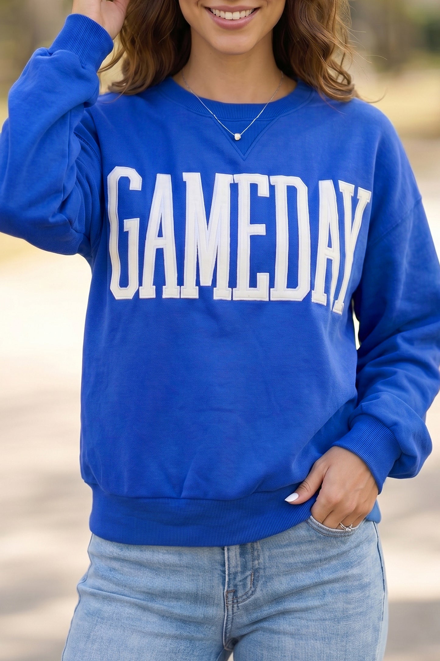 A woman wearing a royal blue crewneck sweatshirt with 'GAMEDAY' printed in large white block letters across the chest, paired with light blue jeans.