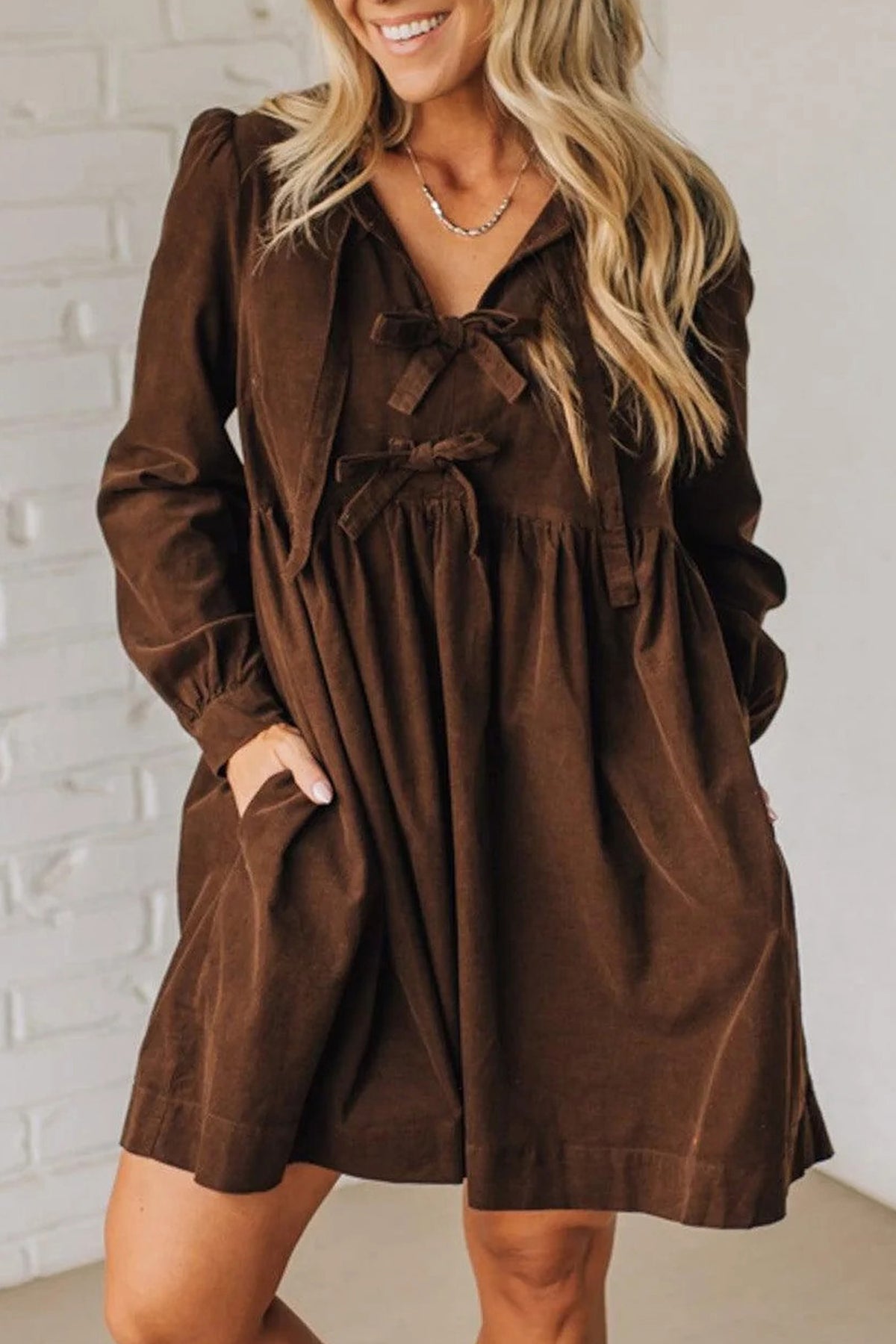 A woman wearing a brown corduroy dress with a tie-neck detail and front pockets, standing against a white brick wall.