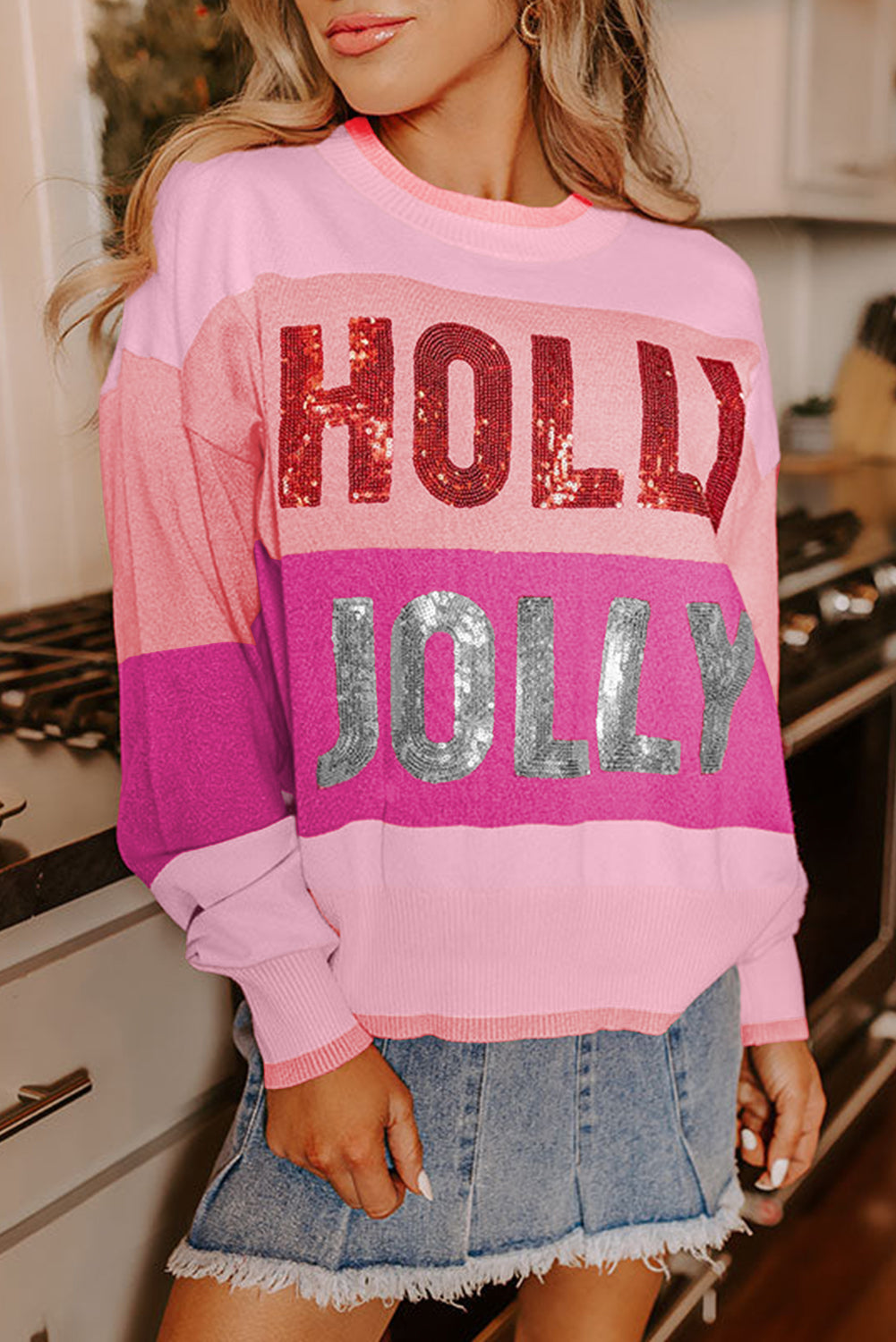 Person wearing a pink sweater with 'Holly Jolly' text in sequins, standing in a kitchen.