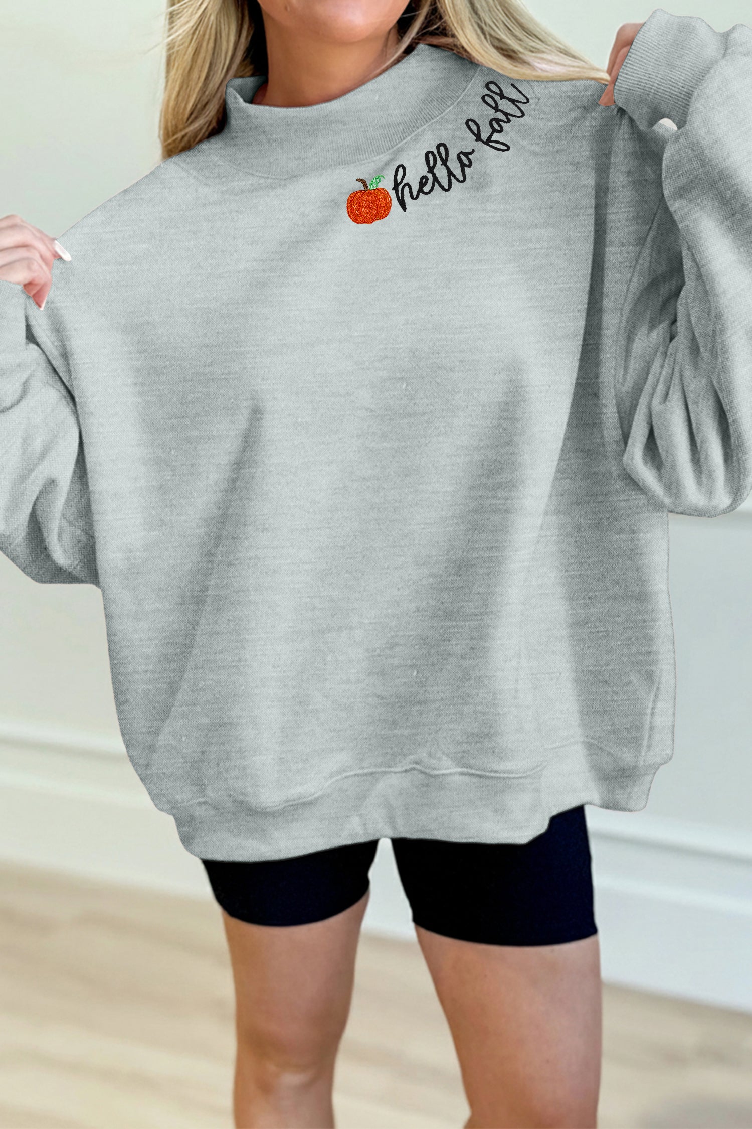 A woman wearing an oversized light gray sweatshirt with a small orange pumpkin graphic and the words 'hello fall' in black script across the chest, paired with black bike shorts.